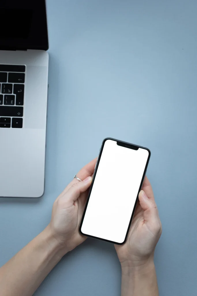 Hands holding a blank-screen smartphone beside a laptop